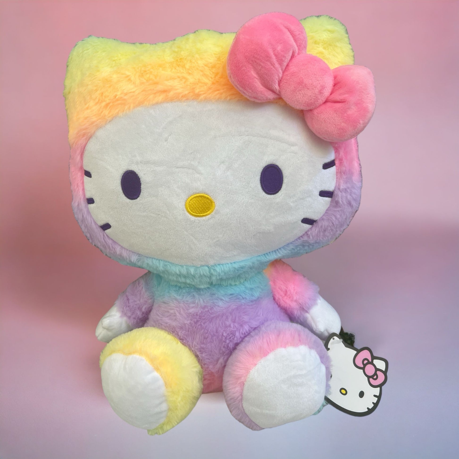 Hello Kitty by Sanrio is wearing a rainbow tie-dye onesie with a pink bow and it's 18 inches tall and it's very soft like a plushie or a pillow